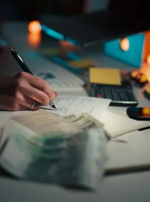 Person writing in a notebook next to stacks of cash and a laptop